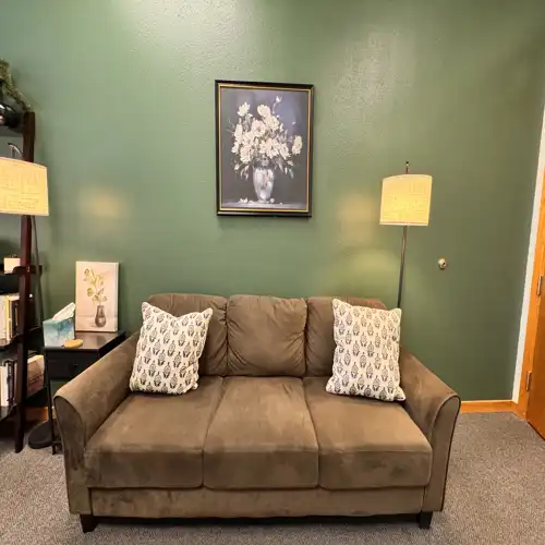 An inviting photo of the therapy space, featuring a plush brown sofa, soft lighting, and a sage green wall, creating a safe and professional atmosphere.