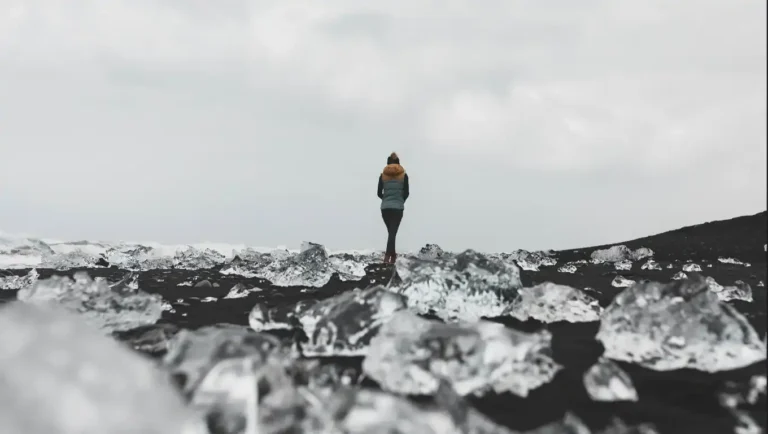 A person walks alone on a black sand beach scattered with chunks of ice.