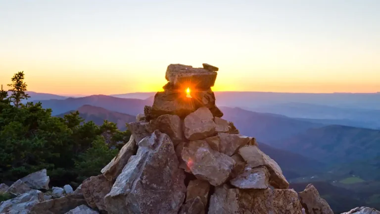 The sun sets behind a mountain range, with its light peeking through a hole in a rock cairn.
