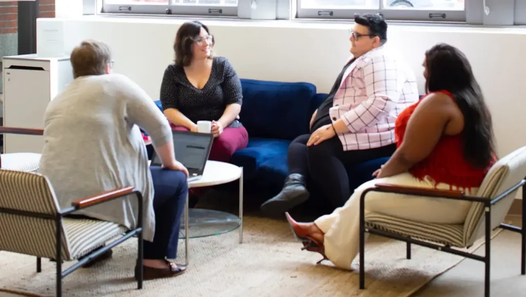 Four people engaged in a relaxed conversation in a comfortable seating area. A supportive community where shared experiences and mutual respect foster healing and growth.