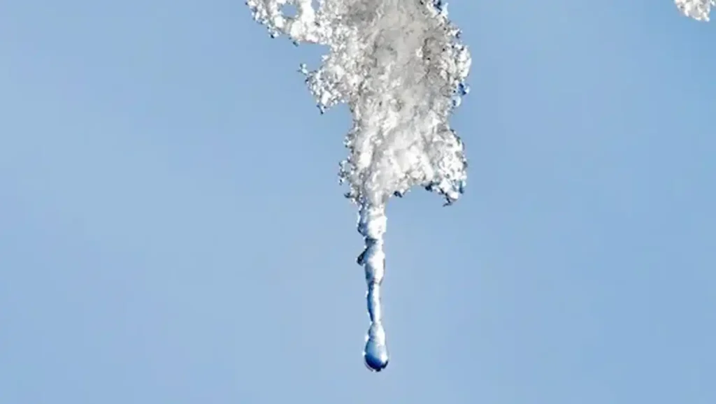 A drop of water falls from a melting icicle against a clear blue sky.