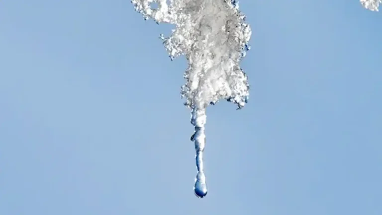 A drop of water falls from a melting icicle against a clear blue sky.