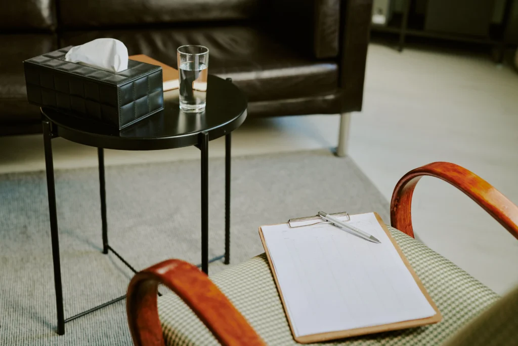 A calming photo of a therapy room setup, which includes a chair with a clipboard, and a small table holding a box of tissues and a glass of water, symbolizing a space prepared for compassionate listening and healing.