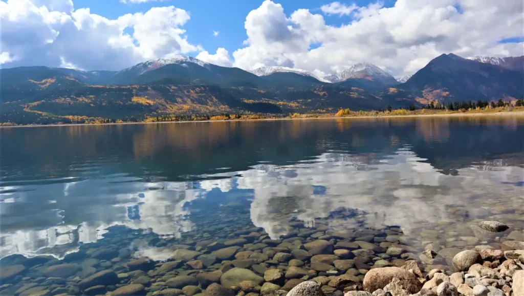 A scenic mountain lake in autumn, with clear water revealing a rocky bottom and snow-capped peaks in the distance.