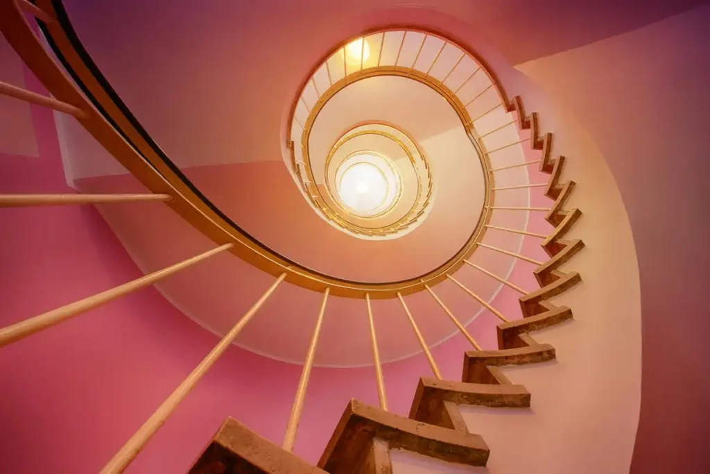 A striking image looking up from the bottom of a winding spiral staircase with pink walls, symbolizing the cyclical nature of obsessions and the upward journey of recovery.