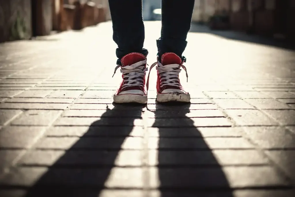 he photo shows the feet and red sneakers of a person standing alone on a sidewalk, with the low sun creating long, dark shadows, which evokes feelings of isolation related to Sexual Addiction.