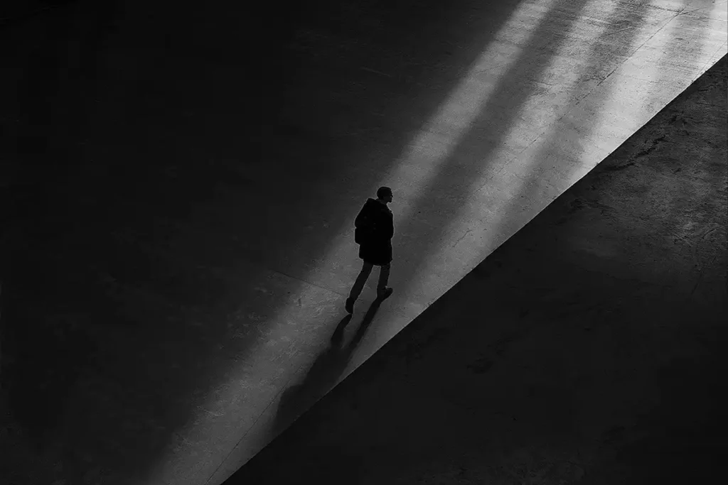 A stark black and white photo of a person walking alone through a sliver of light in a dark space, illustrating an isolating reality of anxiety.