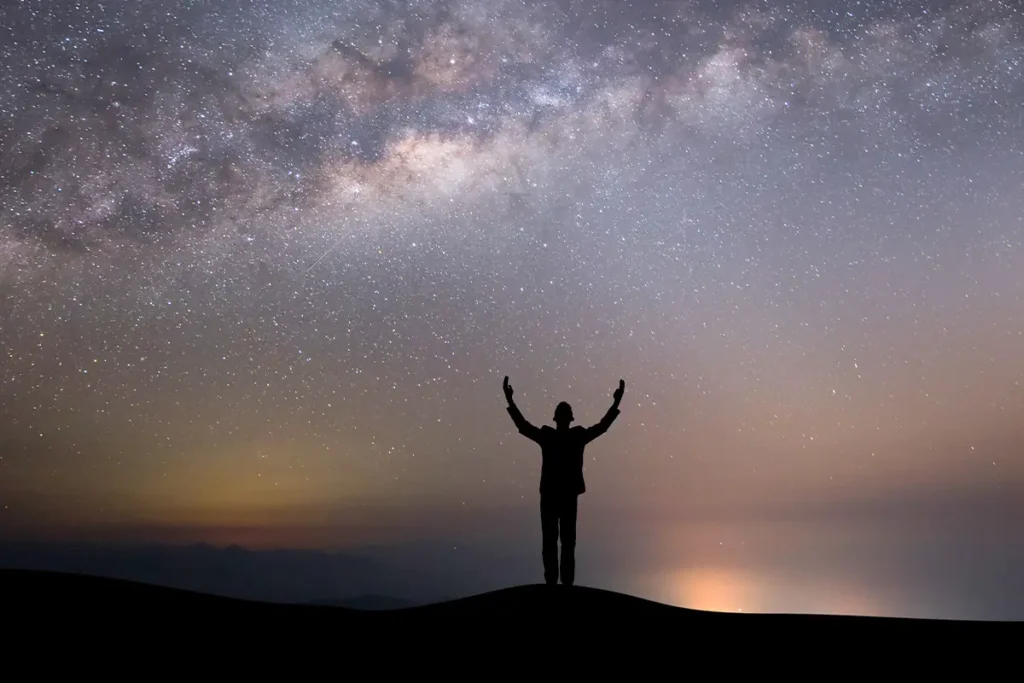 A striking photo of a person's silhouette standing on a hill with arms raised in a gesture of freedom or awe towards a vast, starry night sky with the Milky Way, representing the profound and expansive experience ketamine therapy can offer.