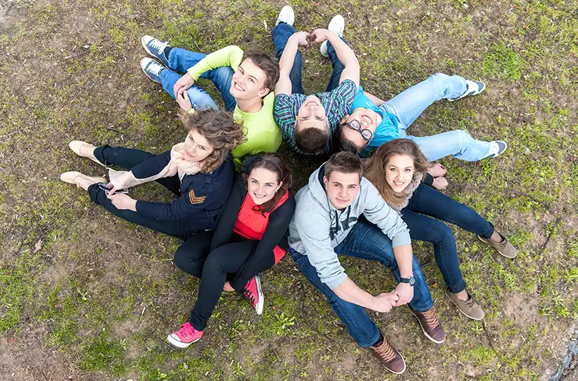 Seven young adults sit in a circle on the grass, looking up at the camera and smiling, symbolizing community and support.