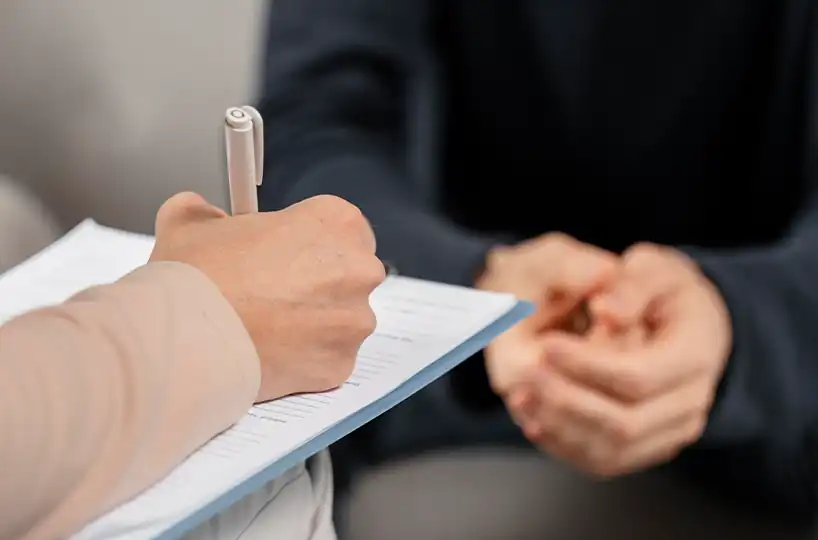 A therapist's hand writing notes on a clipboard during a counseling session, with a client's hands clasped in the blurred background.