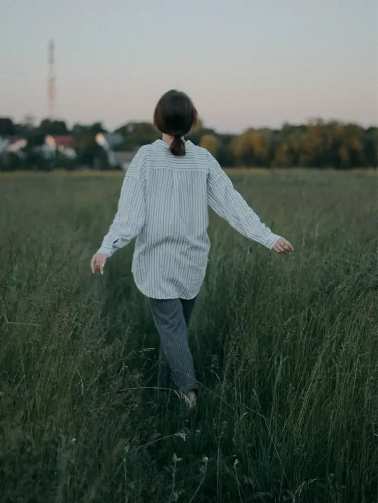 A woman from behind, wearing a striped shirt, walks through a tall grassy field with her arms slightly outstretched, symbolizing freedom and moving forward.