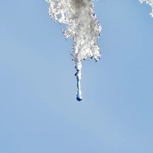 A drop of water falls from a melting icicle against a clear blue sky.