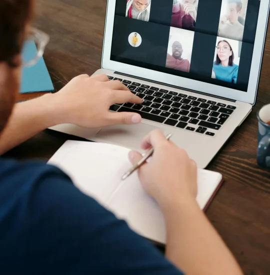 A person taking notes while participating in a virtual group meeting on their laptop.